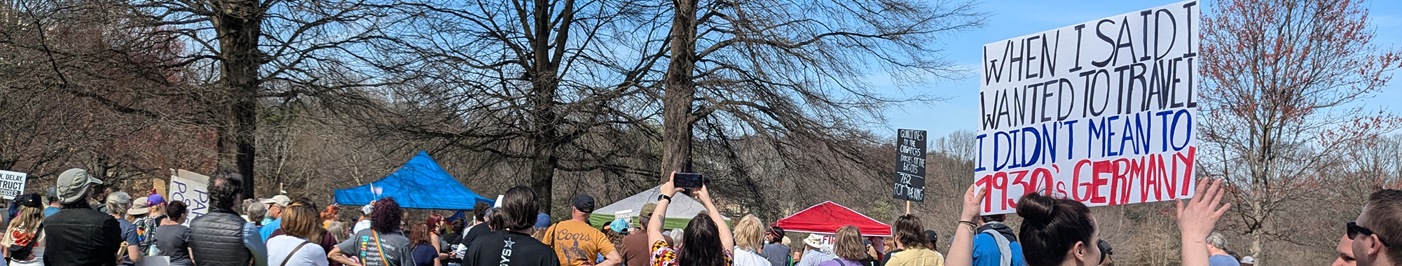 a view of the tops of people's heads at an outdoor rally, a sign reads 'when I said I wanted to travel I didn't mean to 1930s Germany'